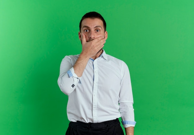 Hombre guapo sorprendido pone la mano en la boca aislada en la pared verde