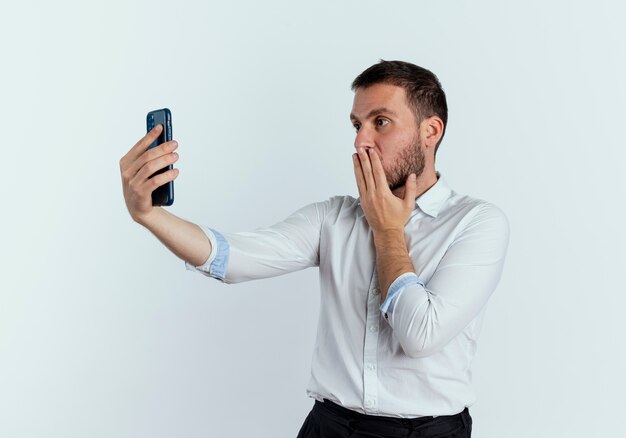 Hombre guapo sorprendido pone la mano en la boca mirando el teléfono aislado en la pared blanca
