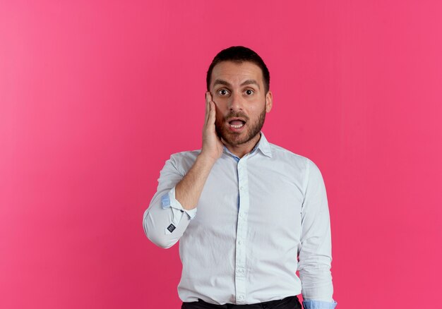 Hombre guapo sorprendido pone la mano en la cara mirando aislado en la pared rosa