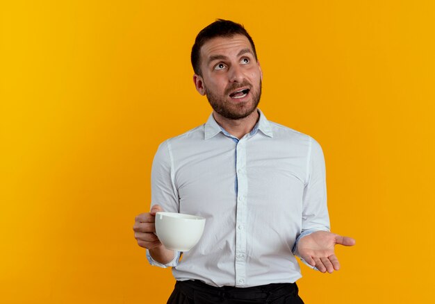 Hombre guapo sorprendido sosteniendo la taza mirando hacia arriba aislado en la pared naranja