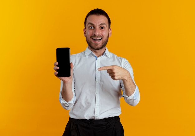 Hombre guapo sorprendido sostiene y apunta al teléfono aislado en la pared naranja