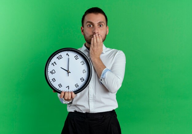 Hombre guapo sorprendido sostiene el reloj y pone la mano en la boca aislada en la pared verde