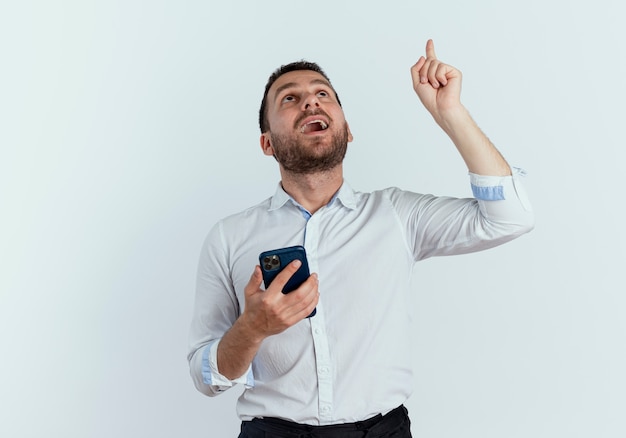 Hombre guapo sorprendido sostiene teléfono mirando y apuntando hacia arriba aislado en la pared blanca