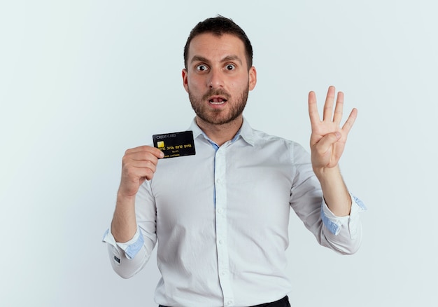 Hombre guapo sorprendido tiene tarjeta de crédito y gestos cuatro con la mano aislada en la pared blanca