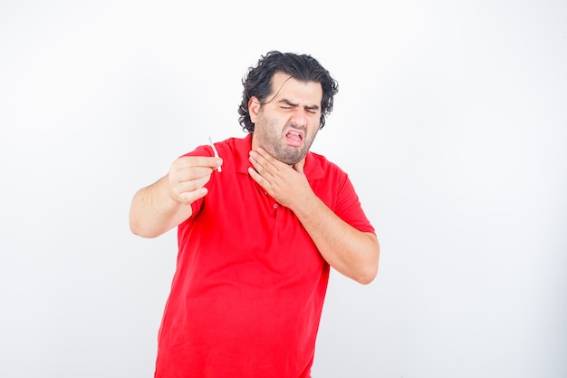 Hombre guapo sosteniendo un cigarrillo, sosteniendo la mano en el cuello, tosiendo con una camiseta roja y mirando disgustado, vista frontal.