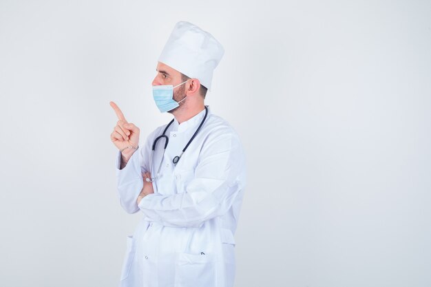 Hombre guapo sosteniendo un estetoscopio alrededor del cuello, apuntando a la izquierda en bata blanca de laboratorio médico, máscara y mirando confiado. vista frontal.