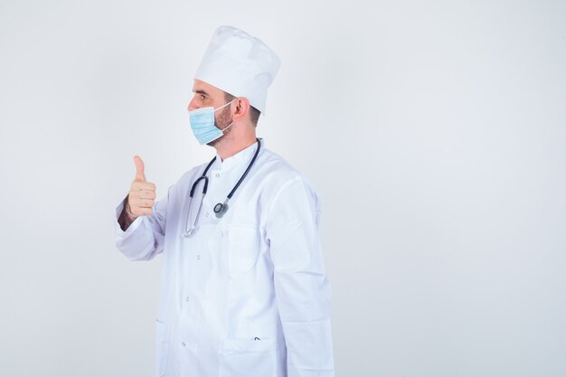 Hombre guapo sosteniendo estetoscopio alrededor del cuello, mostrando el pulgar hacia arriba en bata blanca de laboratorio médico, máscara y mirando confiado, vista frontal.