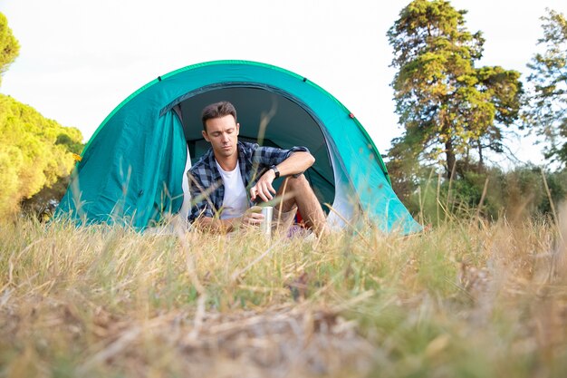 Hombre guapo sosteniendo frasco de vacío con té y sentado en la tienda. Excursionista masculino caucásico relajarse en la naturaleza, disfrutar y acampar en el césped. Turismo de mochilero, aventura y concepto de vacaciones de verano.