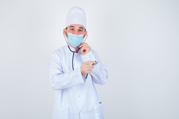 Hombre guapo sosteniendo piezas de estetoscopio en sus oídos como si estuviera escuchando en bata blanca de laboratorio médico, máscara y mirando feliz, vista frontal.