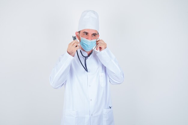 Hombre guapo sosteniendo piezas de estetoscopio en sus oídos como si estuviera escuchando en bata blanca de laboratorio médico, máscara y mirando feliz, vista frontal.