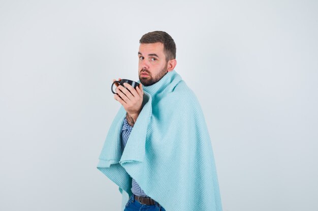 Hombre guapo sosteniendo una taza de té con ambas manos en camisa, jeans, chal y mirando agotado. vista frontal.