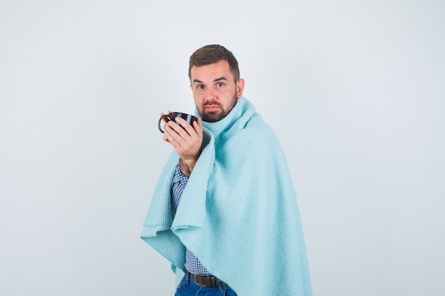 Hombre guapo sosteniendo una taza de té con ambas manos en camisa, jeans, chal y mirando agotado. vista frontal.