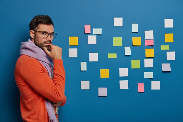 hombre guapo sostiene la barbilla, viste ropa de moda, se para con blocs de notas, mira a través de gafas, piensa en el plan de negocios