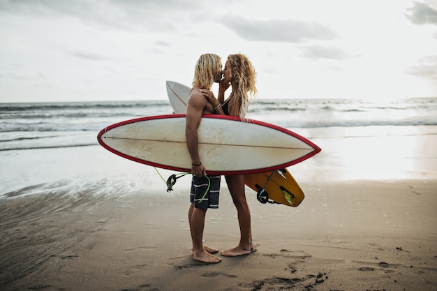 Foto gratuita hombre guapo y su novia se besan contra el fondo del mar y sosteniendo tablas de surf