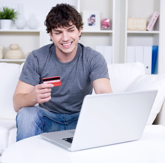 Hombre guapo con tarjeta de crédito y usando laptop para compras en línea - en interiores