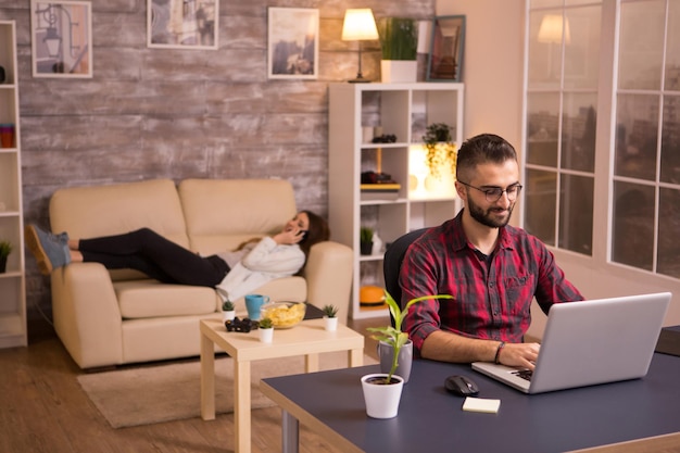 Foto gratuita hombre guapo trabajando en la computadora portátil en la sala de estar y novia relajándose en el sofá hablando por su teléfono. fichas en la mesa.