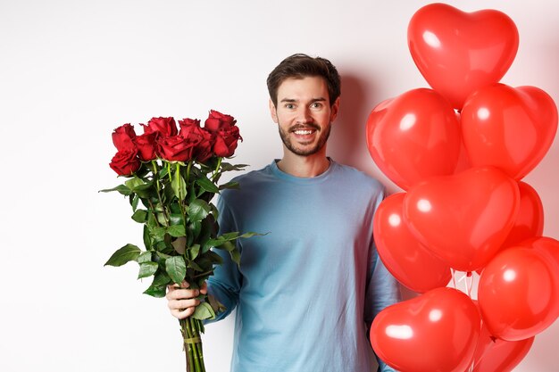 Hombre guapo traer flores y globos de corazones rojos en la fecha del día de San Valentín. Novio romántico con ramo de rosas y regalo para amante, de pie sobre fondo blanco.