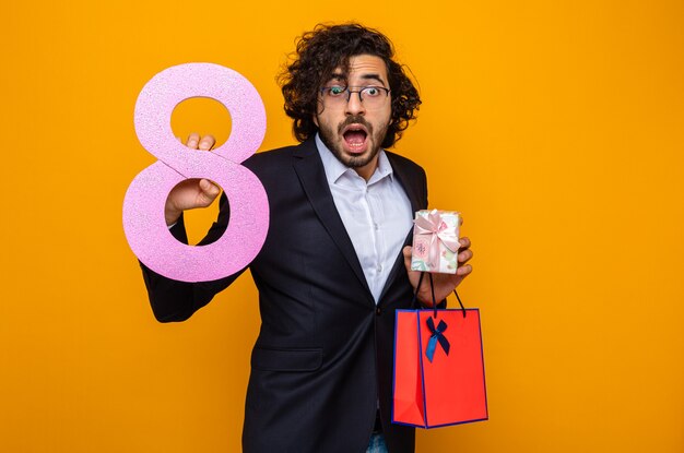 Hombre guapo en traje con bolsa de papel presente con regalo y número ocho mirando a cámara sorprendido y asombrado celebrando el día internacional de la mujer el 8 de marzo de pie sobre fondo naranja