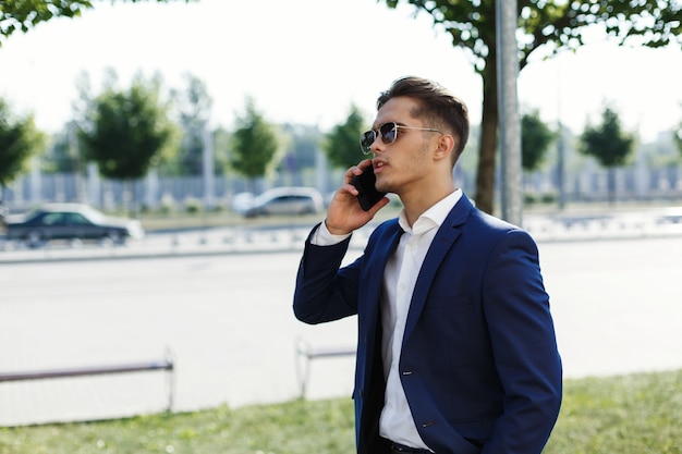 Hombre guapo en un traje camina por la calle en un día soleado y habla en su teléfono inteligente