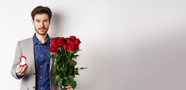 Hombre guapo en traje va a hacer una propuesta de pie con flores de rosas rojas y anillo de compromiso en