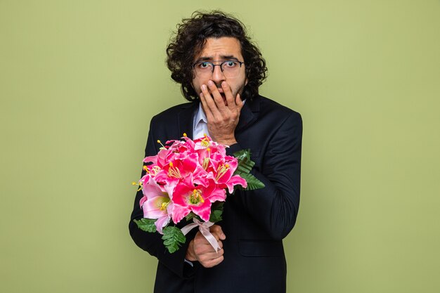 Hombre guapo en traje con ramo de flores mirando sorprendido cubriendo la boca con la mano