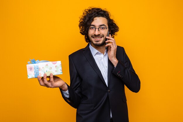 Hombre guapo en traje sosteniendo presente sonriendo alegremente mientras habla por teléfono móvil celebrando el día internacional de la mujer el 8 de marzo de pie sobre fondo naranja