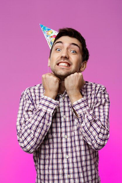 Hombre guapo tratando de sacar el sombrero de cumpleaños sobre pared púrpura.