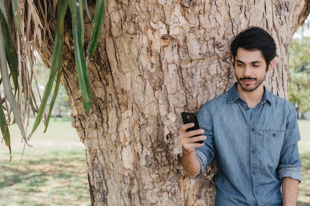 Hombre guapo viendo el teléfono debajo de un árbol