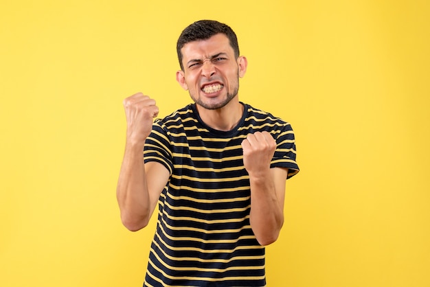 Hombre guapo de vista frontal en camiseta a rayas blanco y negro que muestra gesto ganador sobre fondo amarillo aislado