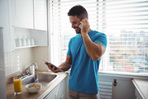 Hombre hablando por teléfono móvil mientras usa tableta digital en la cocina