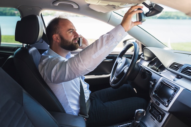 Hombre hablando por teléfono móvil que ajusta el espejo retrovisor del coche