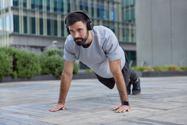 El hombre hace ejercicio de tabla demuestra poses de resistencia en urbano escucha música en auriculares hace ejercicios de fuerza entrena cuerpo hace flexiones