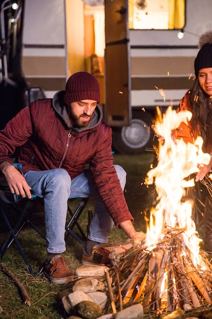 Hombre haciendo más fuerte el fuego del campamento en una fría noche de otoño en las montañas. Turistas con autocaravana retro.