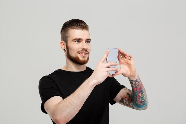 Hombre hermoso joven que mira el teléfono sobre la pared gris.