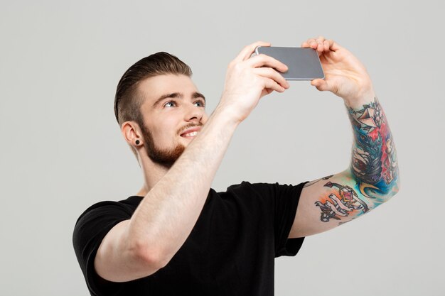 Hombre hermoso joven que mira el teléfono sobre la pared gris.