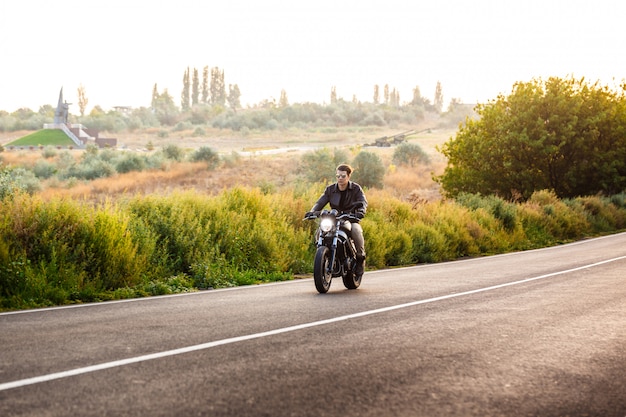 Hombre hermoso joven que monta en la moto en el camino del campo.