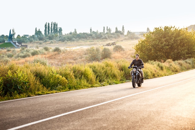 Hombre hermoso joven que monta en la moto en el camino del campo.