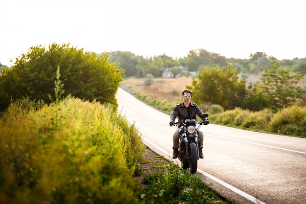 Hombre hermoso joven que monta en la moto en el camino del campo.