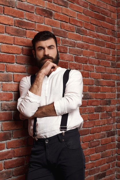 Hombre hermoso joven que piensa posando en la pared de ladrillo.