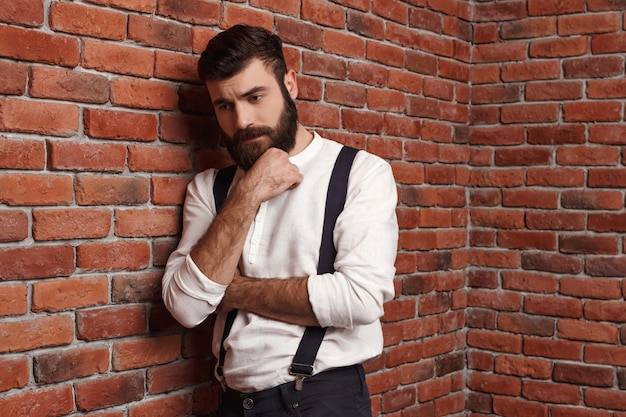 Hombre hermoso joven que piensa posando en la pared de ladrillo.