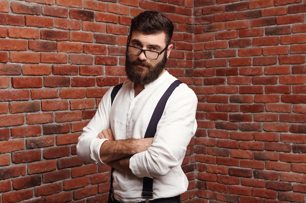 Foto gratuita hombre hermoso joven que presenta con los brazos cruzados en la pared de ladrillo.