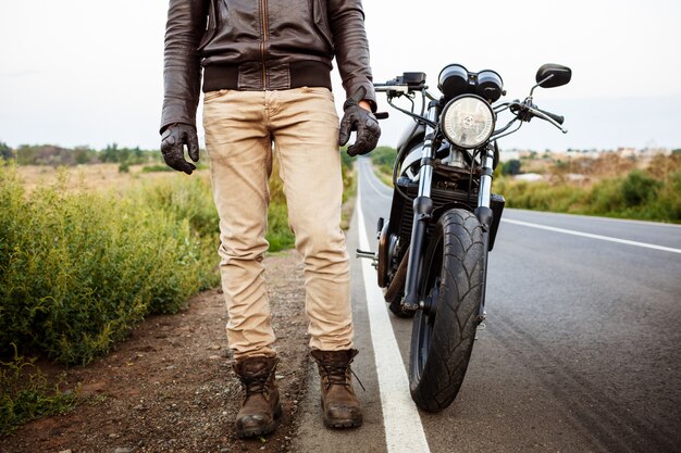 Hombre hermoso joven que presenta cerca de su moto en el camino del campo.