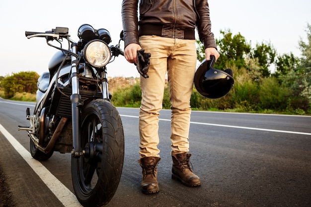 Hombre hermoso joven que presenta cerca de su moto en el camino del campo.