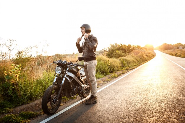 Hombre hermoso joven que presenta cerca de su moto en el camino del campo.