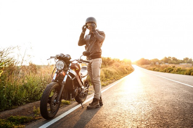 Hombre hermoso joven que presenta cerca de su moto en el camino del campo.