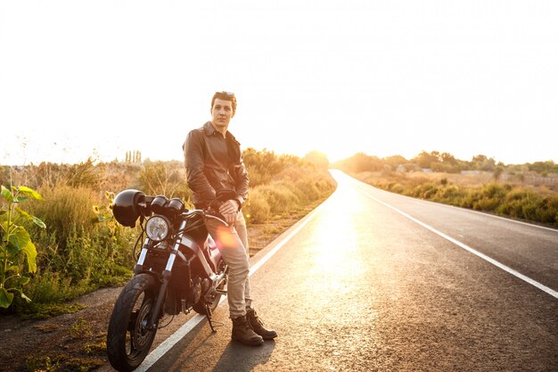 Hombre hermoso joven que presenta cerca de su moto en el camino del campo.