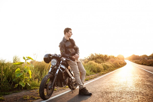 Hombre hermoso joven que presenta cerca de su moto en el camino del campo.