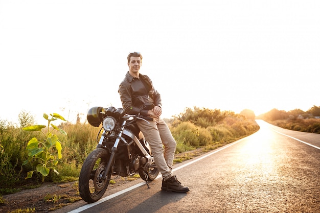 Hombre hermoso joven que presenta cerca de su moto en el camino del campo.