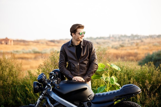 Foto gratuita hombre hermoso joven que presenta cerca de su moto en el camino del campo.