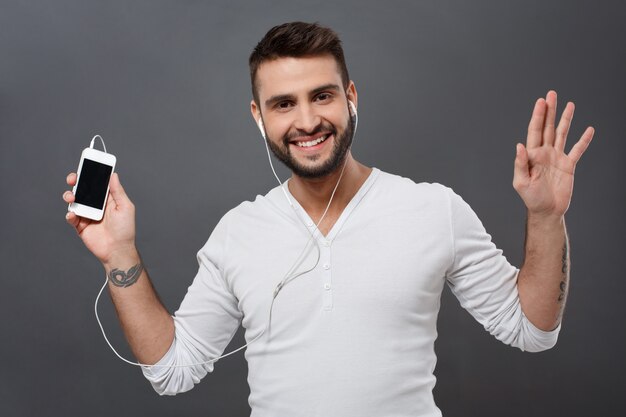 Hombre hermoso joven que sonríe sosteniendo el teléfono sobre la pared gris